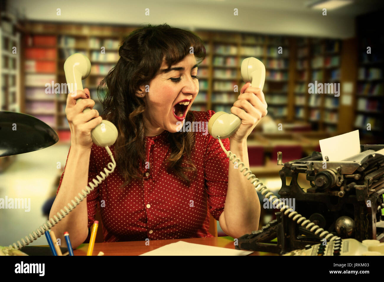 Jeune femme avec robe rouge désespérément avec deux téléphones à deux mains dans Office, 1960 Banque D'Images