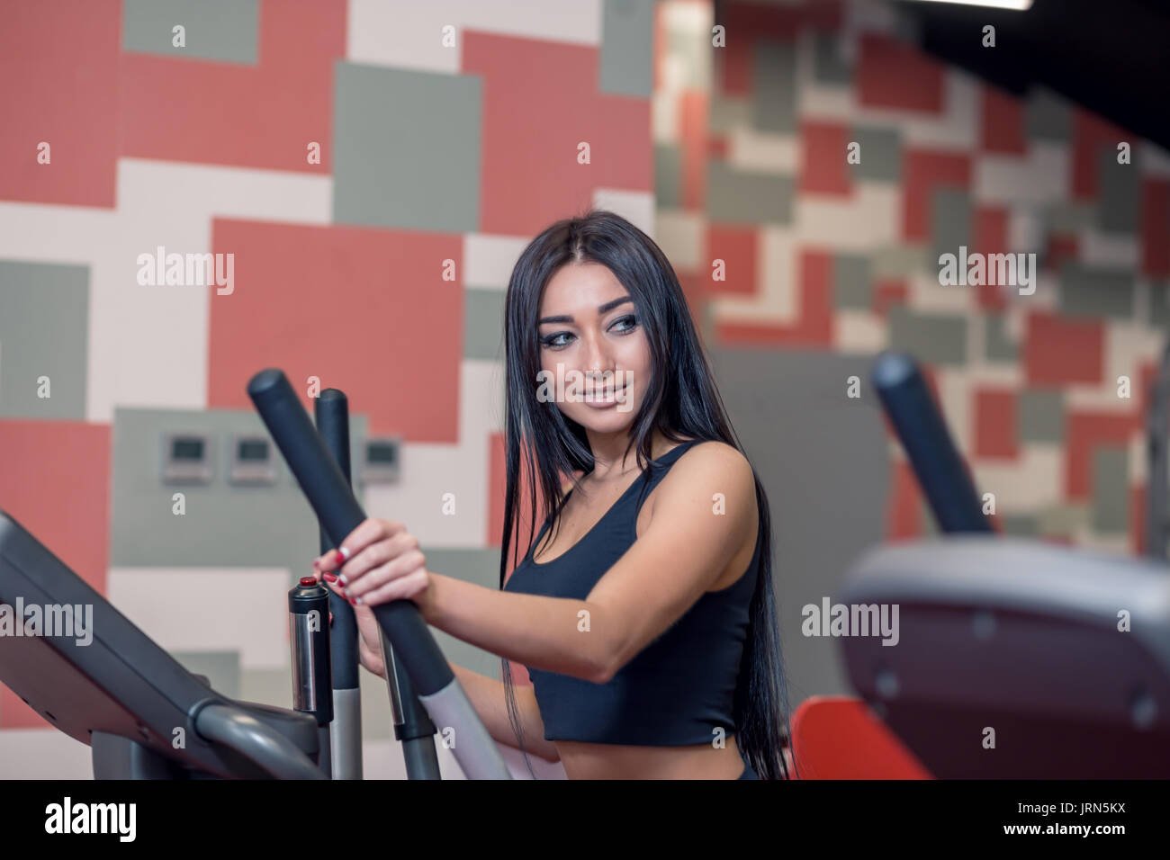 Portrait de belle femme brune sportive exerçant à l'aide d'une machine elliptique suivant pour mettre en place l'homme et souriant pour lui au cours de l'exercice dans la salle de sport moderne Banque D'Images