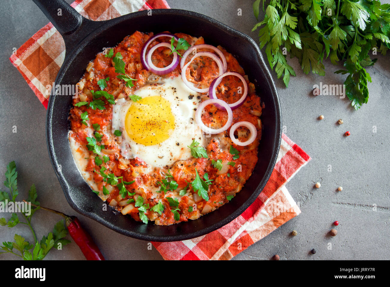 Shakshuka dans une poêle en fonte. Style moyen-orientale oeufs pochés dans une sauce tomate et du poivre avec le persil, l'oignon et les épices sur fond de béton gris. Banque D'Images