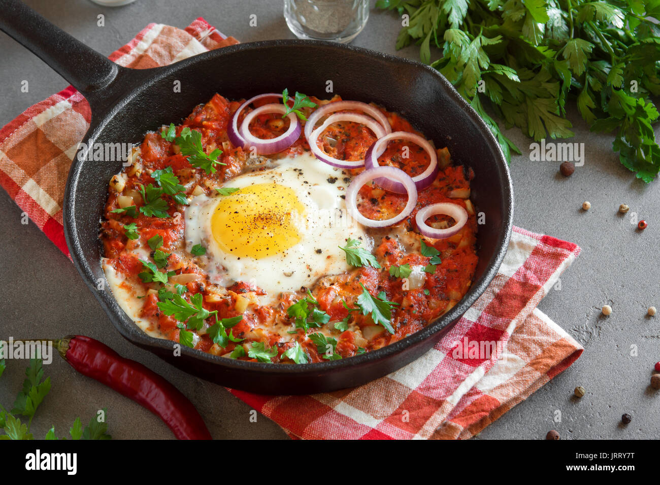 Shakshuka dans une poêle en fonte. Style moyen-orientale oeufs pochés dans une sauce tomate et du poivre avec le persil, l'oignon et les épices sur fond de béton gris. Banque D'Images