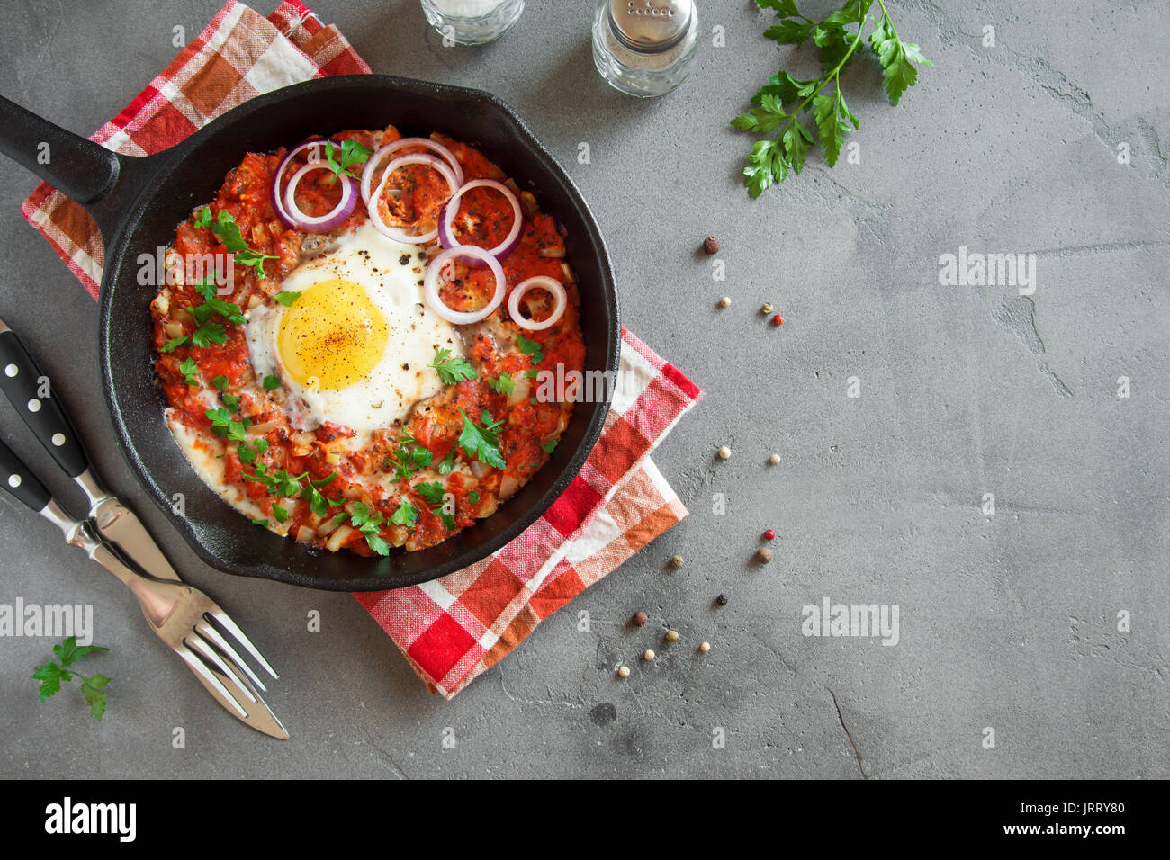 Shakshuka dans une poêle en fonte. Style moyen-orientale oeufs pochés dans une sauce tomate et du poivre avec le persil, l'oignon et les épices sur fond de béton gris. Banque D'Images