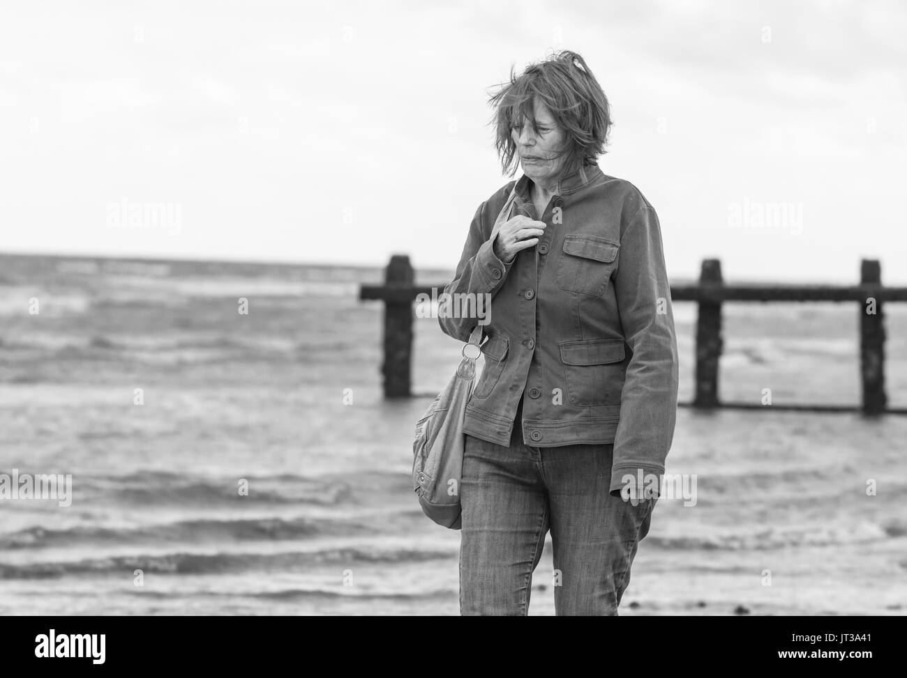 Personnes âgées / femme d'âge moyen à pied par la mer sur la côte par un jour de vent au Royaume-Uni. Noir et blanc. Banque D'Images