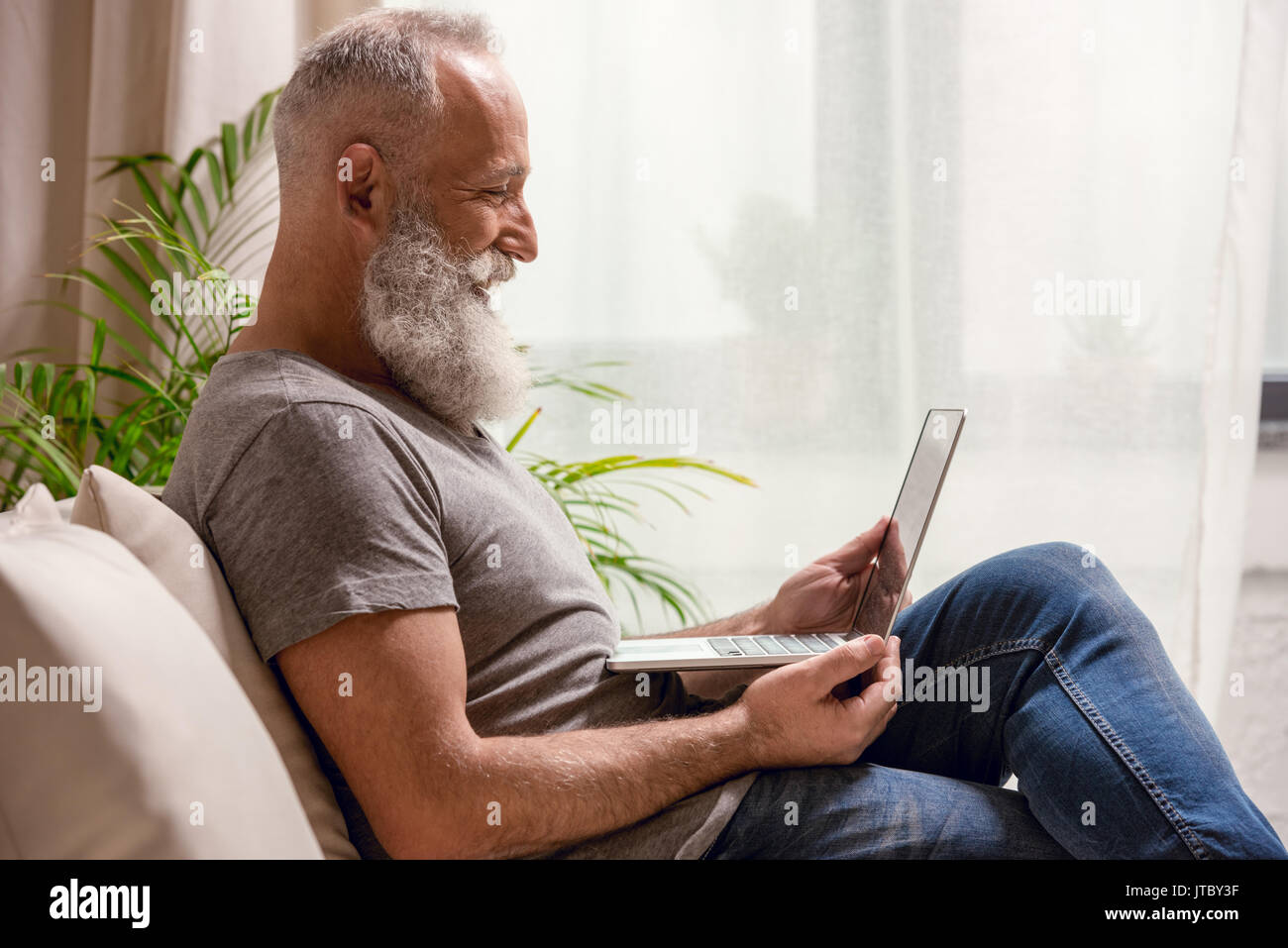 Homme barbu senior smiling and looking at laptop while sitting on sofa at home Banque D'Images