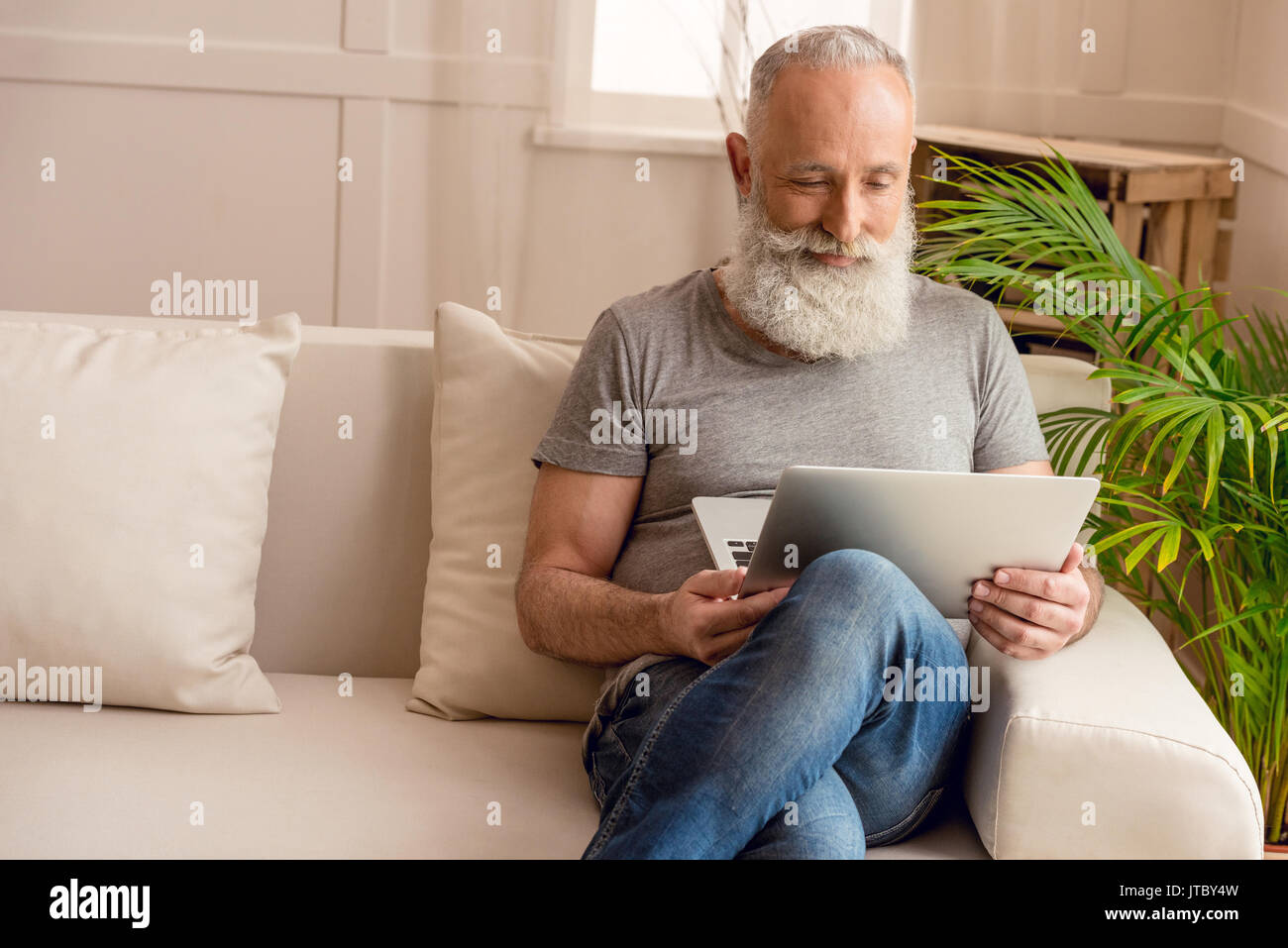 Happy senior homme barbu en utilisant laptop while sitting on sofa at home Banque D'Images