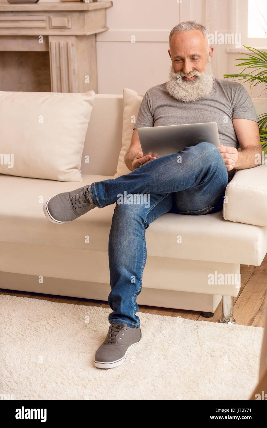 Happy senior homme barbu en utilisant laptop while sitting on sofa at home Banque D'Images