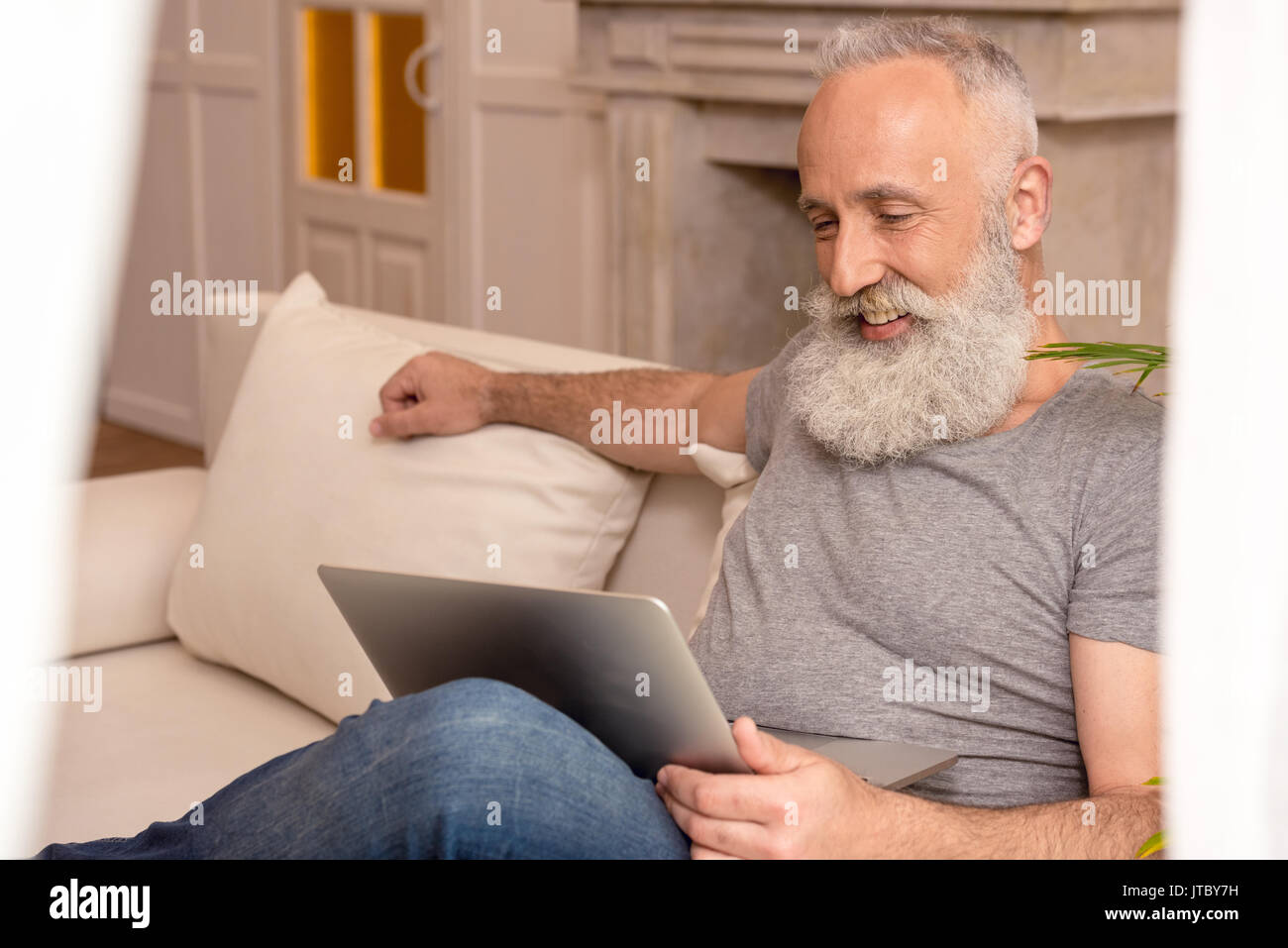 Happy senior homme barbu en utilisant laptop while sitting on sofa at home Banque D'Images