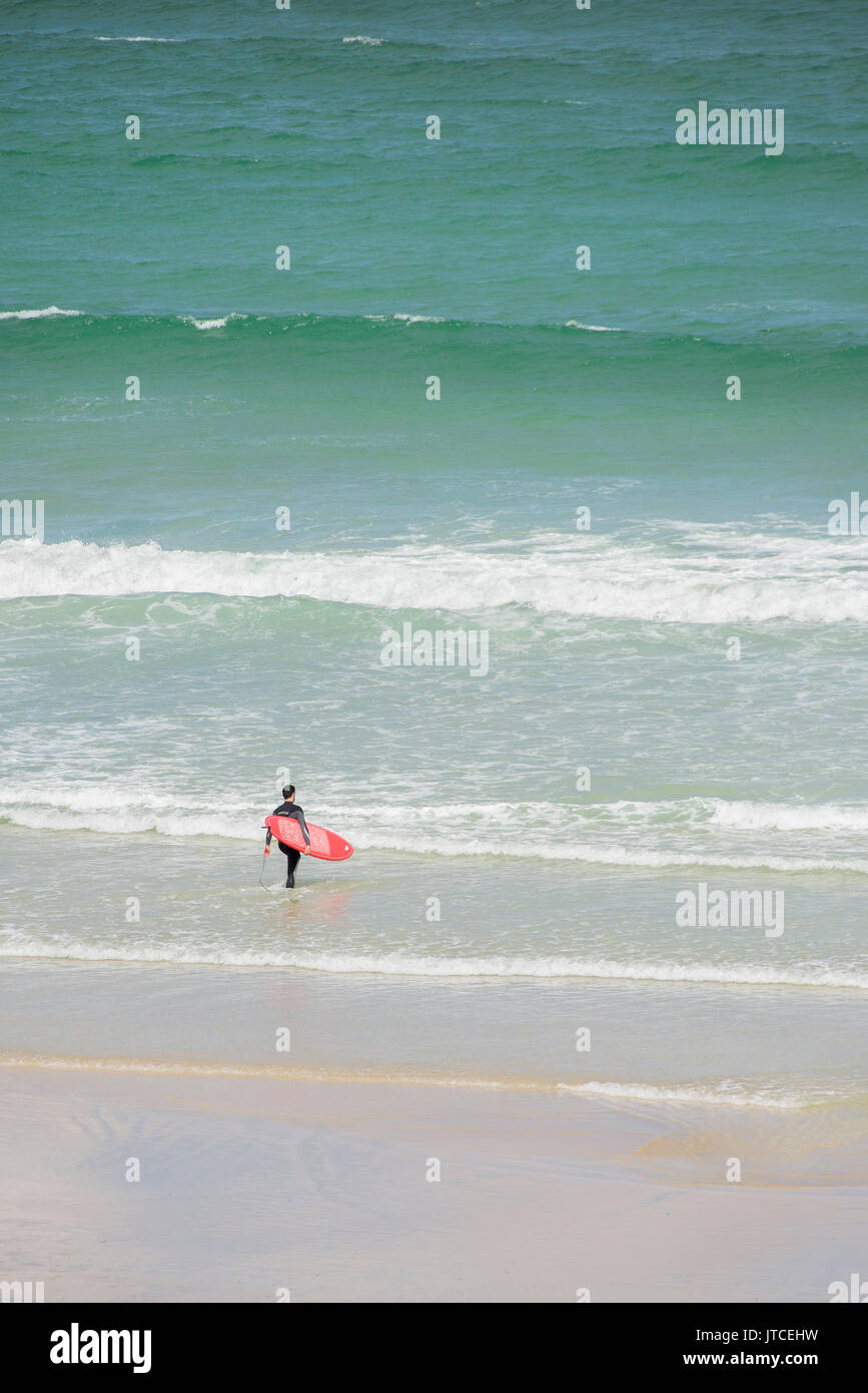 Un surfeur solitaire avec une planche rouge marcher dans la mer. Newquay, Cornwall. Banque D'Images