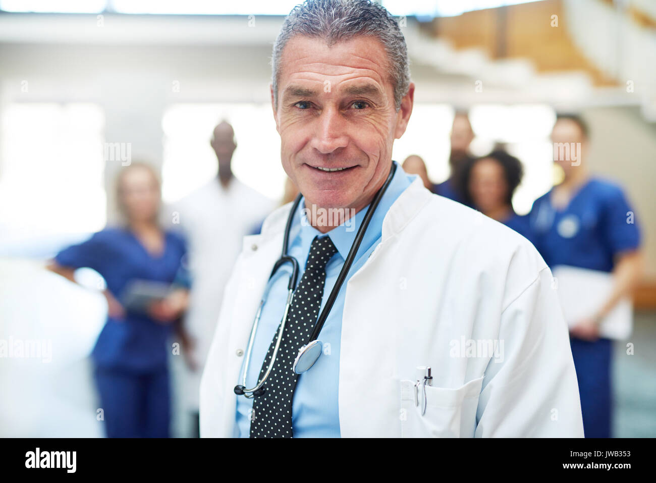 Mature positif permanent médecin dans une clinique et à la recherche à l'appareil photo. Comité permanent de l'équipe médicale en arrière-plan Banque D'Images