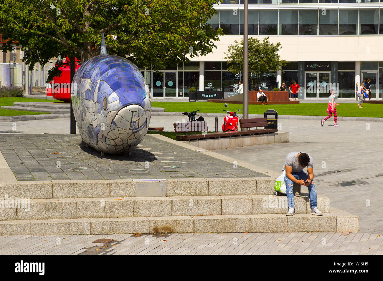 Un jeune homme assis à côté de texter le gros poisson sculpture sur le Donegall Quay dans la zone du port de Belfast, en Irlande du Nord Banque D'Images