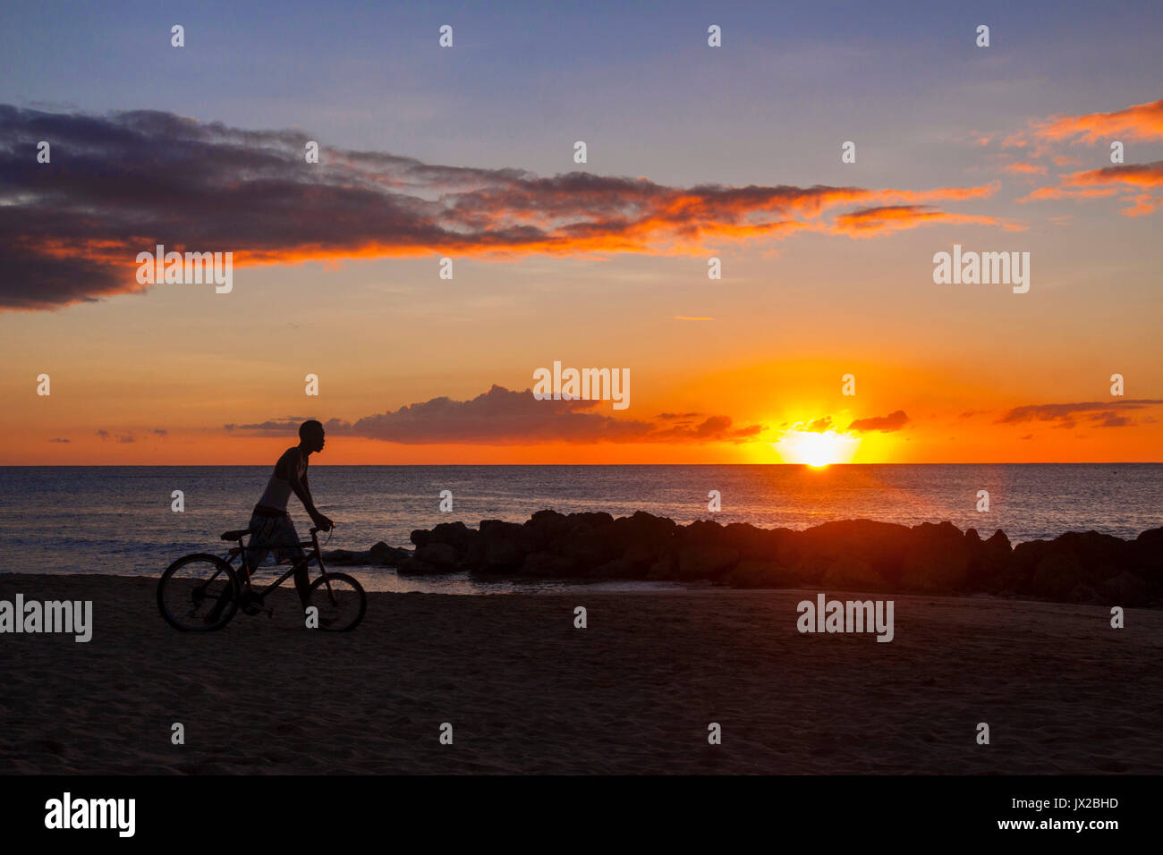 Un jeune mam pousse sa bicyclette le long de la plage au coucher du soleil à Port St Charles, la Barbade Banque D'Images