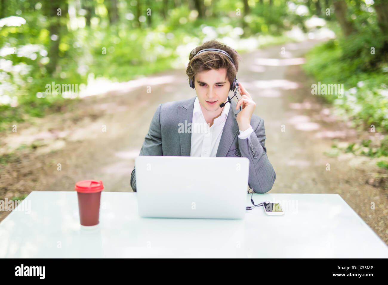 Handsome Businessman with headset sitting au bureau bureau avec ordinateur portable et tasse de café au centre d'appels à Green Forest Park . Banque D'Images