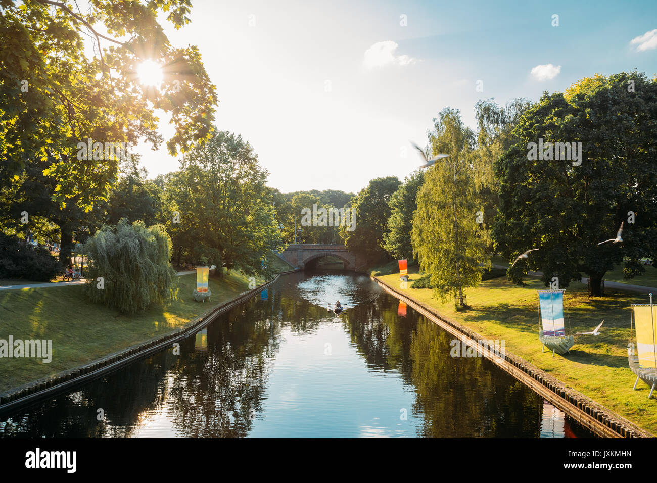 Riga, Lettonie. Ville Rivière Canal dans le parc de la colline Bastion. Soleil qui brille à travers les feuilles vertes sous le soleil de soir d'été. Fontaines dans l'eau de rivière Canal. Banque D'Images