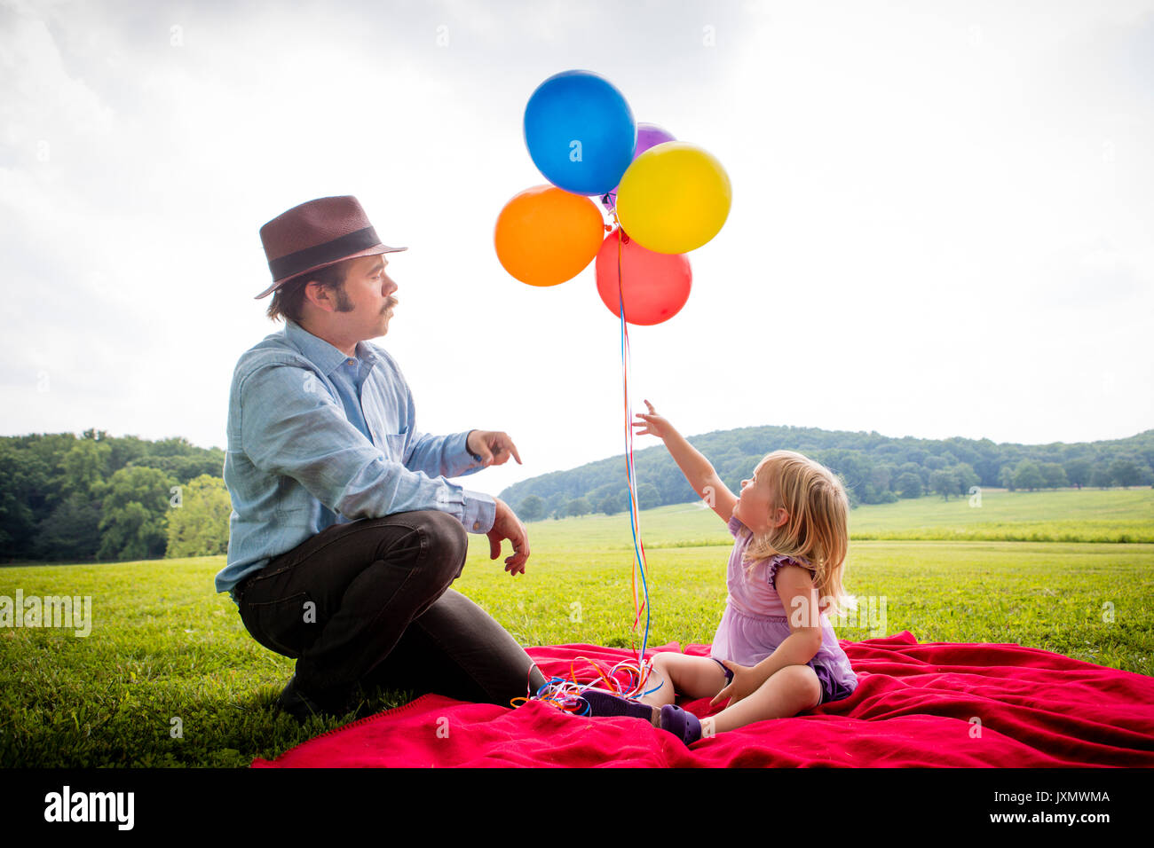 Fille assise avec père pointant vers le haut pour bouquet de ballons colorés in rural field Banque D'Images