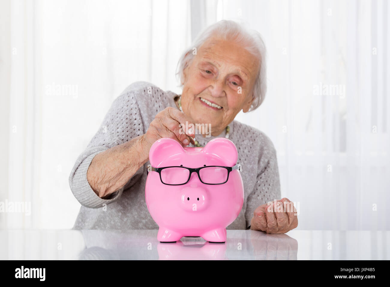 Happy Senior Woman Inserting Coin In Piggybank On Table Banque D'Images