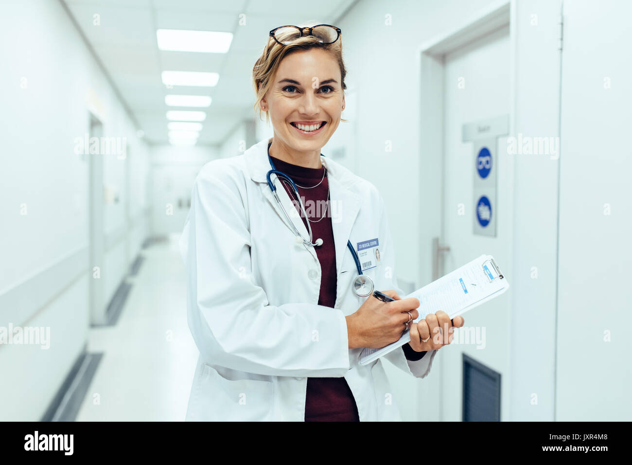 Portrait of happy young female doctor standing in hospital corridor et l'écriture de rapport médical sur le presse-papiers. Caucasian woman travaillant en soins infirmiers. Banque D'Images