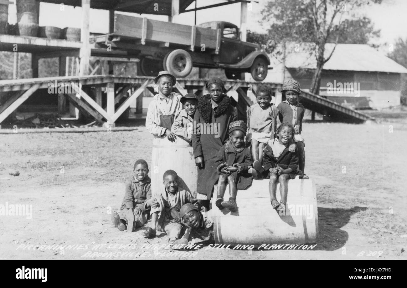 Portrait d'un groupe de jeunes Afro-américains, quelques enfants debout ou assis sur un tonneau, de petits enfants assis sur la terre, Et les enfants plus âgés debout, souriant expressions du visage, devant une grande rampe en bois avec un camion sur elle, sur la plantation Lewis à Brooksville, Floride, 1930. Banque D'Images
