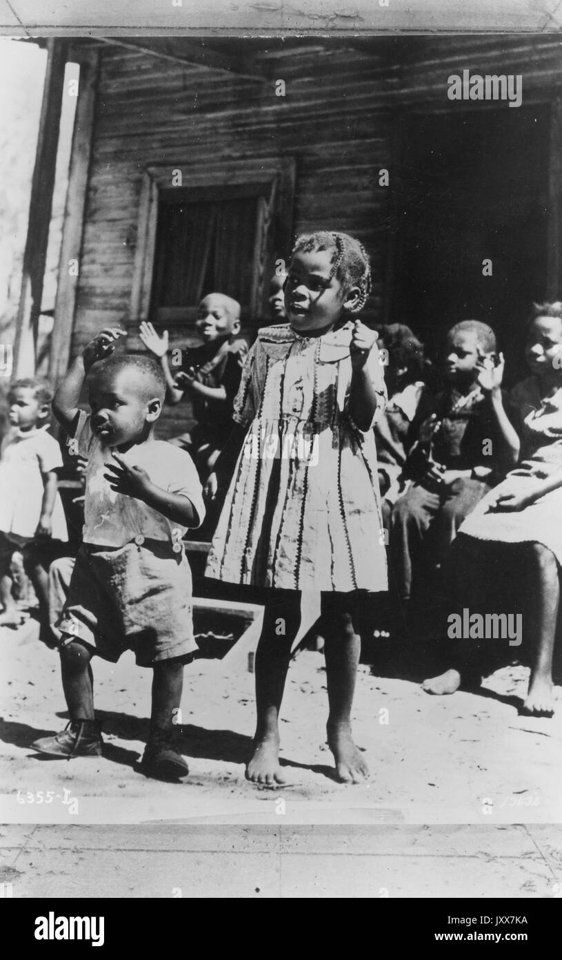 Portrait d'enfants afro-américains, dansant et claquant devant un porche, deux à l'avant, un garçon avec sa main au-dessus de sa tête et l'autre sur sa poitrine, et une fille pieds nus dans une robe sombre, sur la plantation Lewis à Brooksville, Floride, 1930. Banque D'Images
