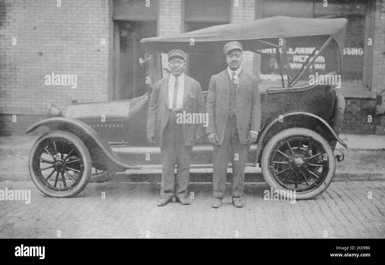 Deux hommes afro-américains portant des costumes et des chapeaux devant une voiture, devant un magasin de nettoyage à sec, expressions faciales neutres, 1920. Banque D'Images