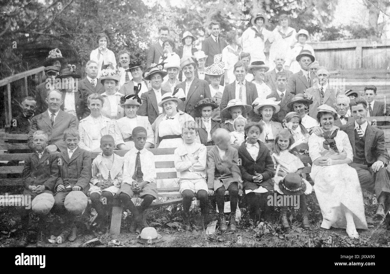 Photo de paysage pleine longueur d'adultes et d'enfants, certains assis sur un banc, d'autres debout, à l'extérieur, tous habillés formellement, expressions du visage souriantes, 1910. Banque D'Images