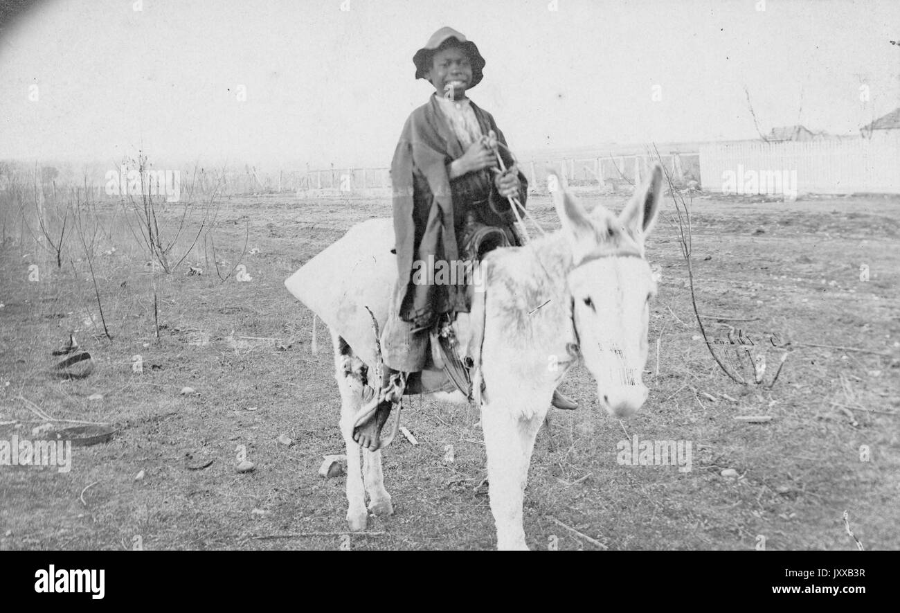Portrait assis de jeunes Afro-américains sur poney, portant une chemise claire, un pantalon clair, un châle foncé et un chapeau foncé, assis dans un champ clairsemé, expression souriante, 1915. Banque D'Images