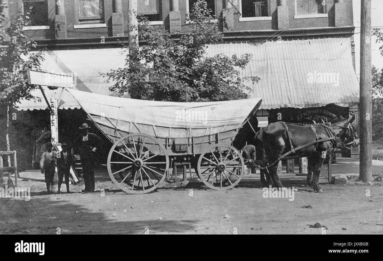Un homme afro-américain avec deux enfants afro-américains debout à côté d'un chariot tiré par des chevaux, portant des vêtements et des chapeaux sombres, debout devant le magasin et des arbres sur la piste de terre, 1910. Banque D'Images