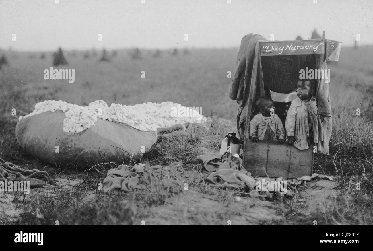 Deux jeunes enfants afro-américains portant des vêtements légers debout dans une structure en bois marquée « Day nursery » avec des couvertures, debout à côté d'une grosse pile de coton récolté, se tenant debout sur le terrain, expressions neutres, 1915. Banque D'Images