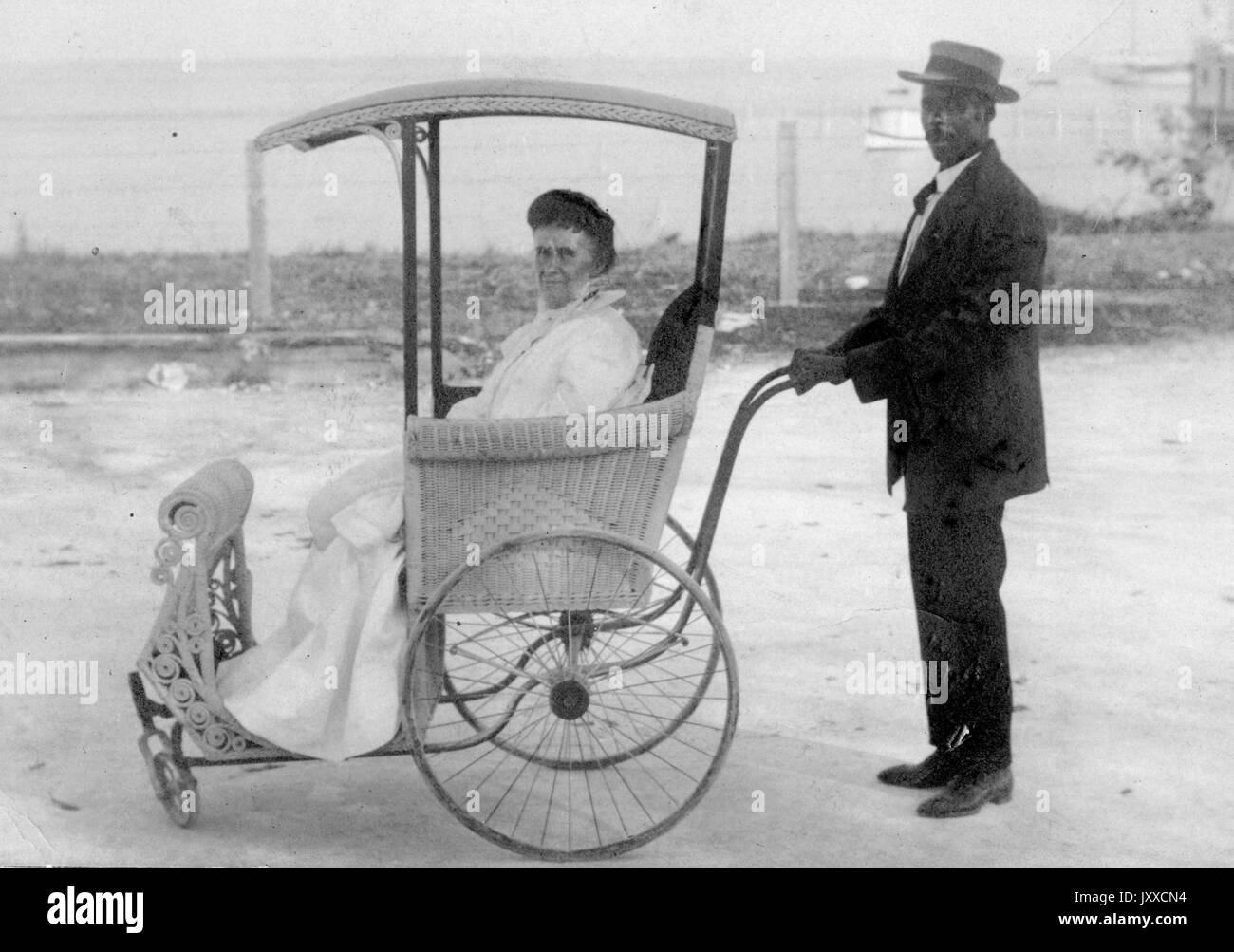 Portrait complet d'un homme afro-américain portant un costume et un chapeau sombres, poussant une femme blanche mûre qui porte une robe blanche dans un chariot avec de grandes roues, 1920. Banque D'Images
