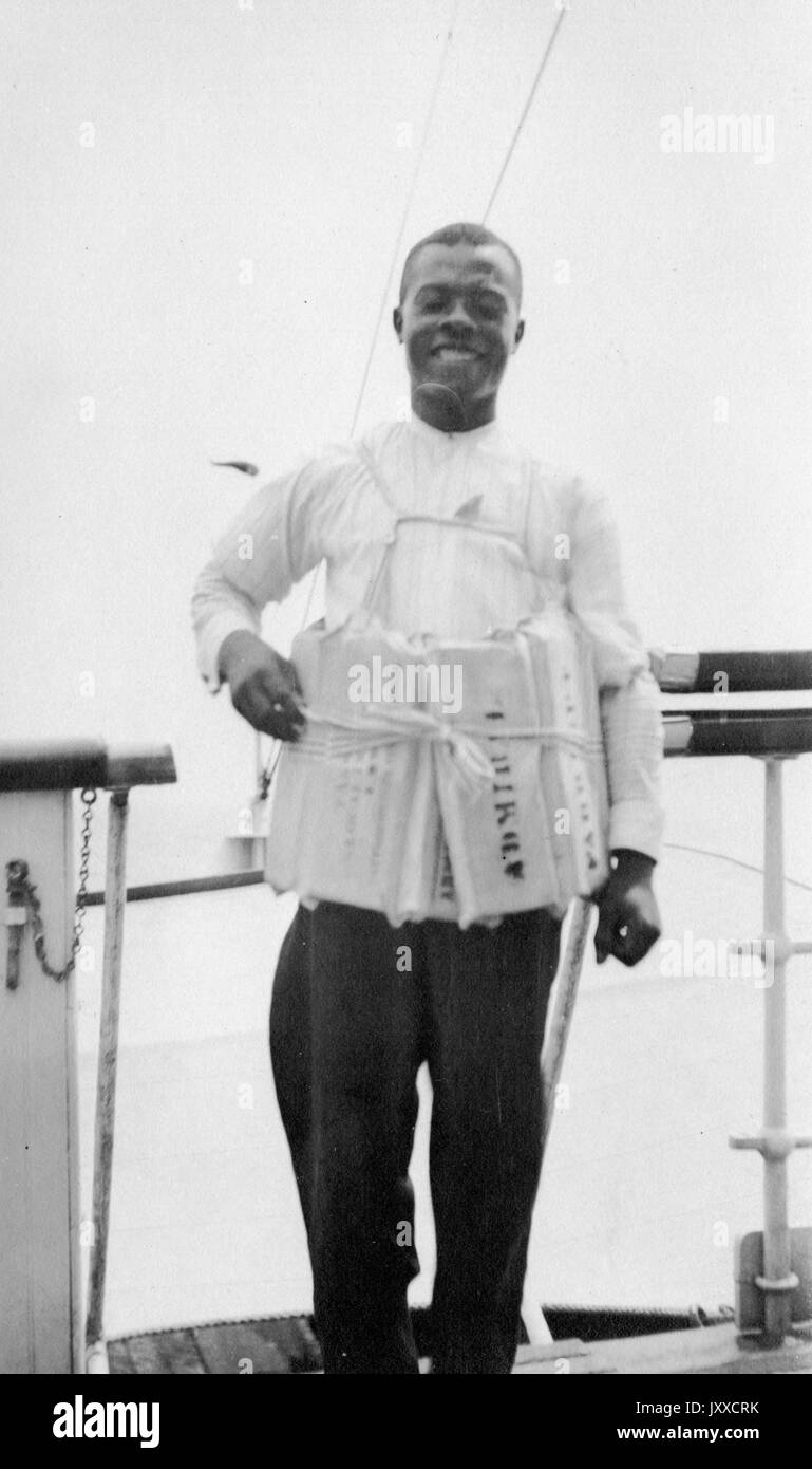 Trois quarts de portrait d'un homme afro-américain debout, avec des parchements attachés autour de son corps, avec une expression faciale souriante, portant une chemise blanche et un pantalon sombre, 1920. Banque D'Images