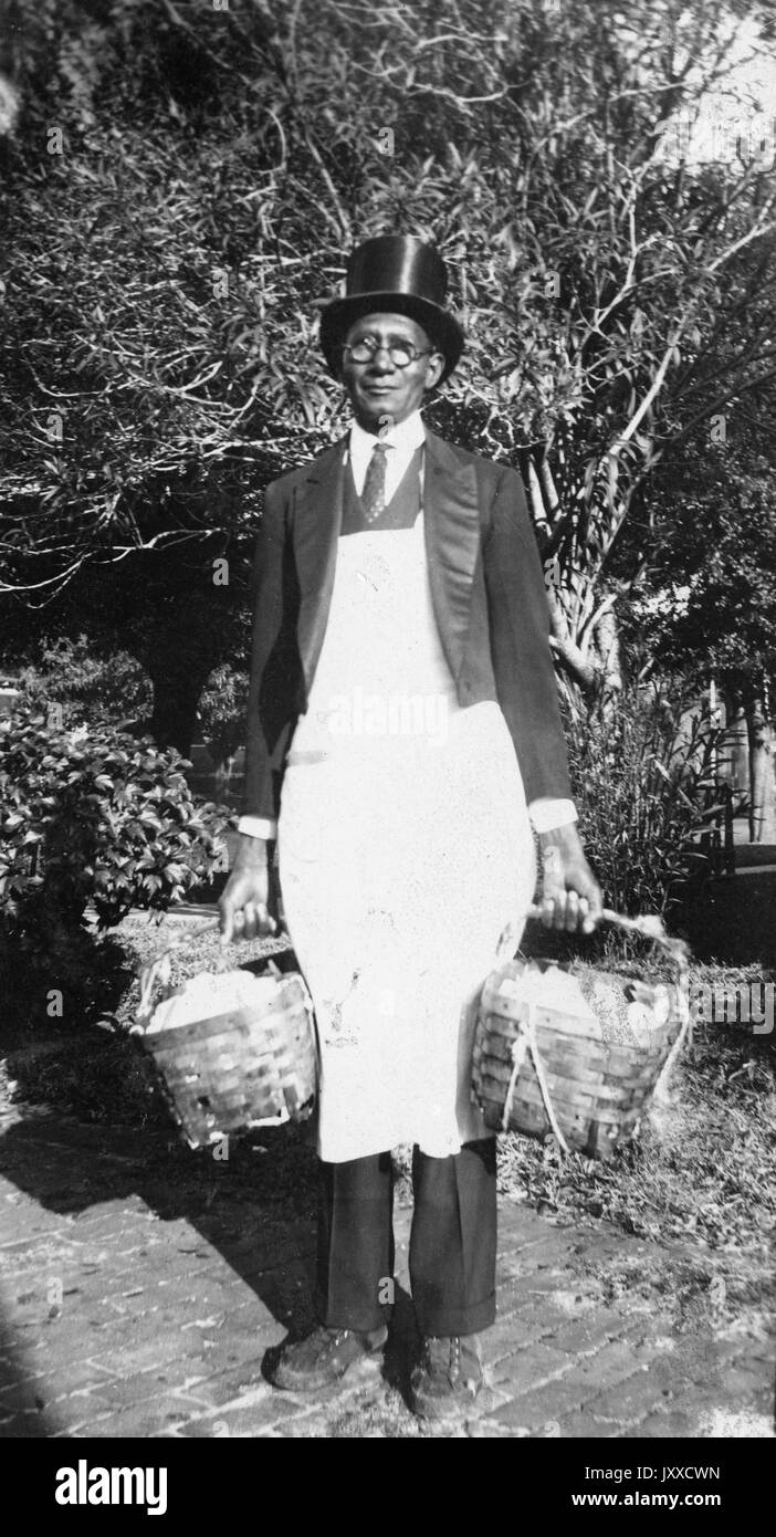 Portrait debout sur toute la longueur d'un homme afro-américain mature, avec une expression neutre sur un chemin de briques devant un arbusté, portant un panier dans l'une ou l'autre main, portant un chapeau, des lunettes, un costume, une cravate et un tablier, 1915. Banque D'Images