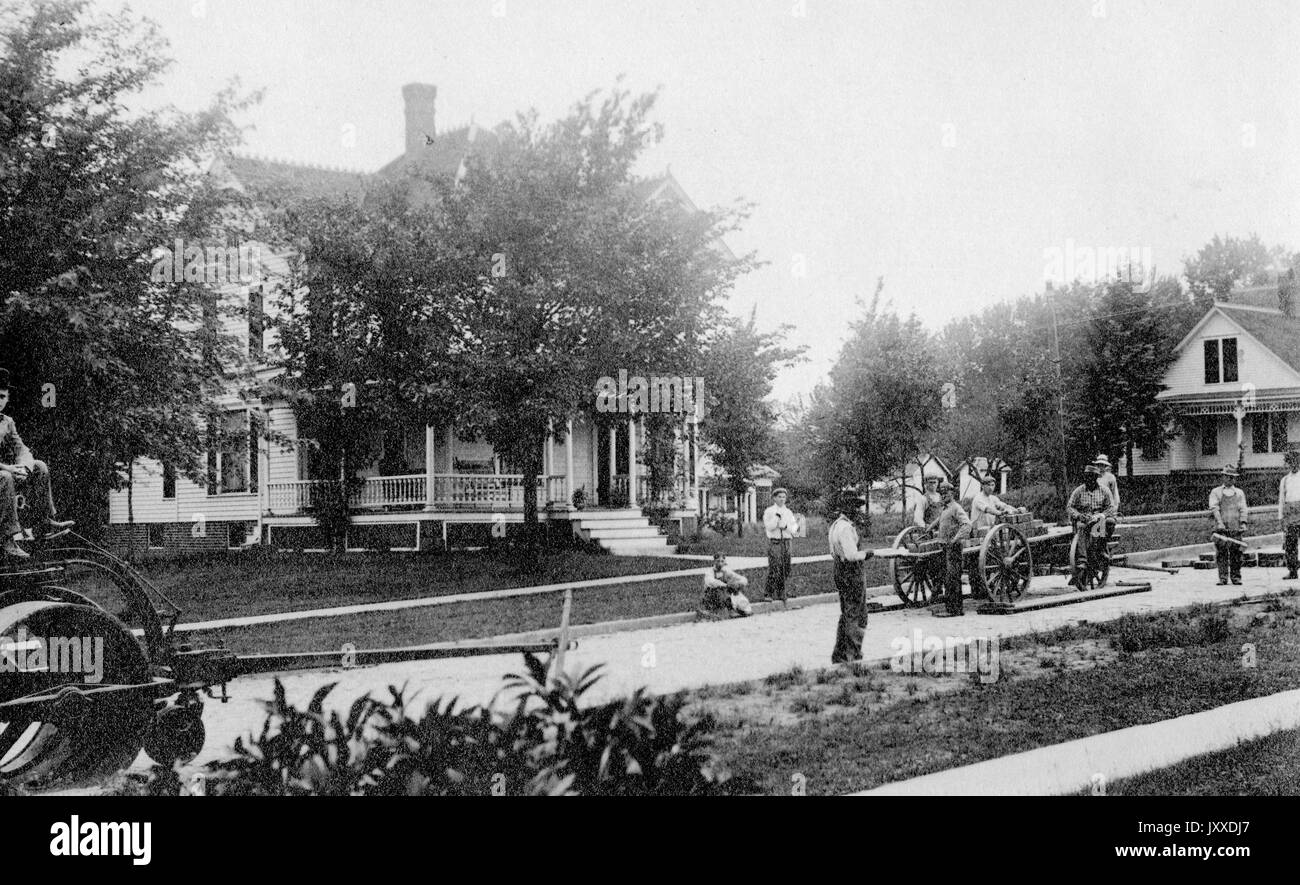 Un groupe de travailleurs américains pose un chemin, en utilisant un petit wagon ouvert pour transporter des pavés, devant les maisons dans une ville américaine, 1925. Banque D'Images