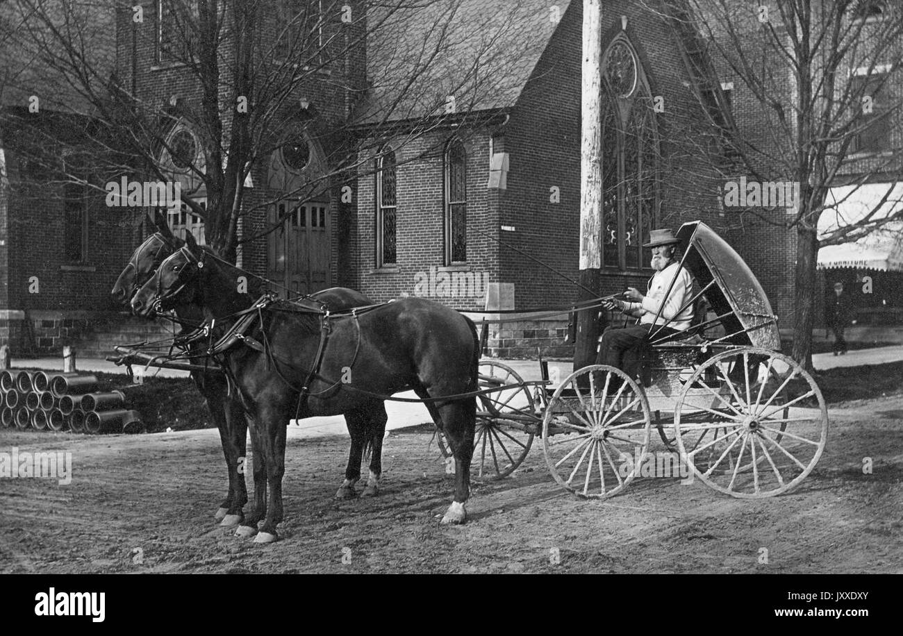 Un ouvrier africain américain mature, portant un chapeau, une chemise légère et un pantalon sombre, conduit un petit wagon, transporté par deux chevaux, à l'extérieur de ce qui semble être une église, 1920. Banque D'Images