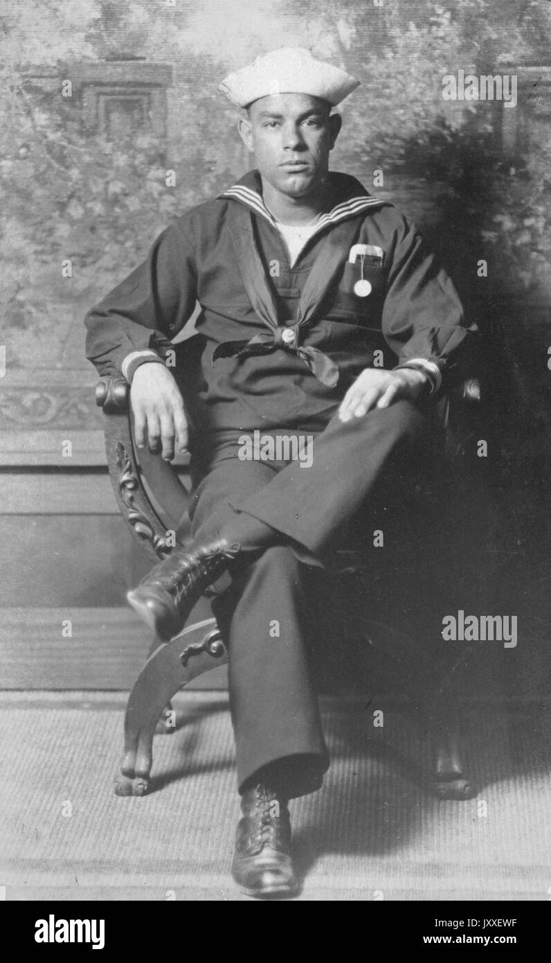 Portrait d'un marin de la marine américaine africaine assis dans une chaise sculptée en bois devant une murale de fleurs, il porte un uniforme de Sailor foncé et un chapeau de Sailor de couleur claire, ses bras reposent sur les bras de la chaise et une cheville est franchie au-dessus de l'autre jambe au genou, 1920. Banque D'Images