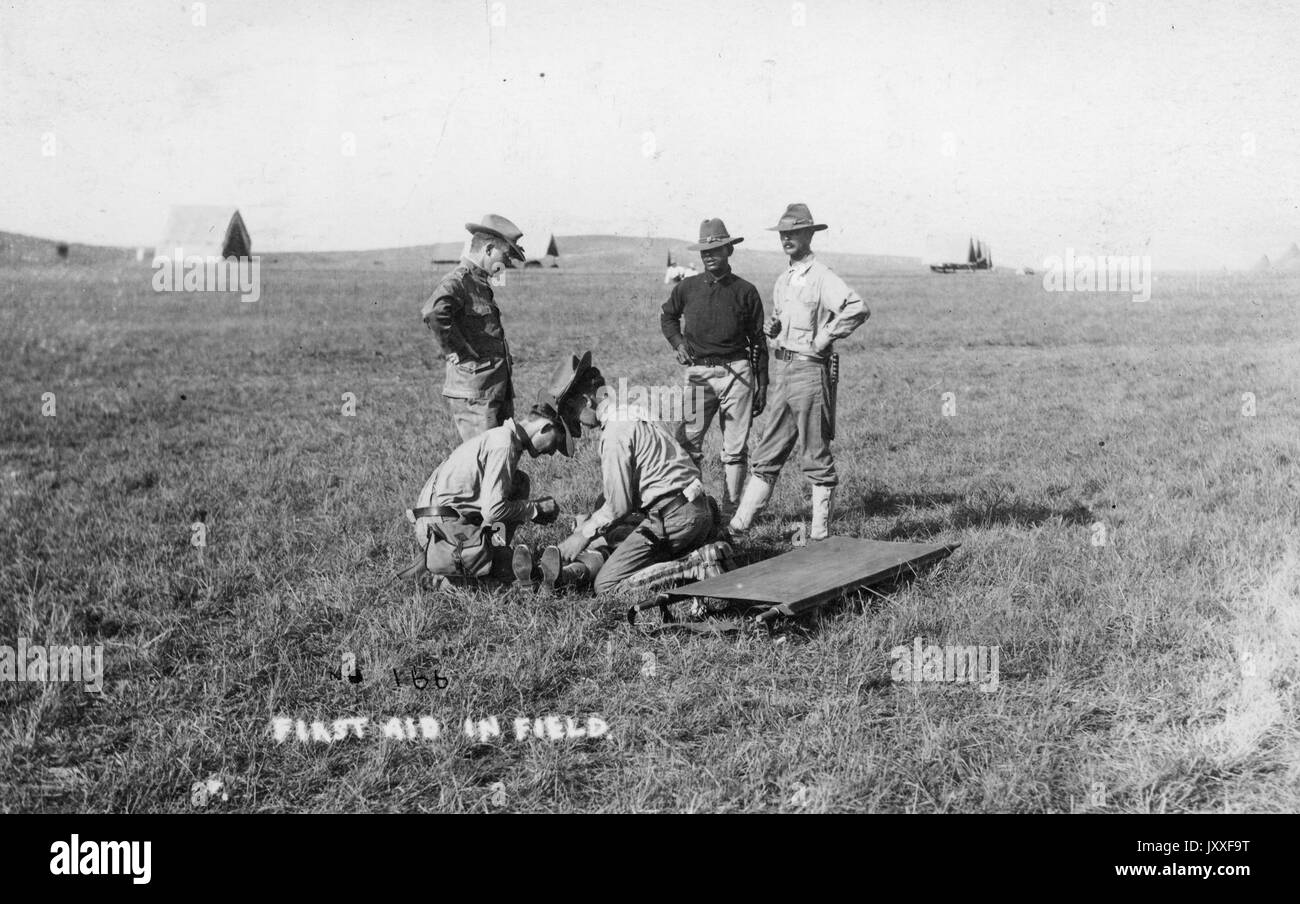 Un groupe de travailleurs sont au milieu d'un champ avec trois tentes militaires tout au fond, deux des hommes se penchent sur le terrain de l'examen d'un homme allongé sur le sol tandis que trois hommes, l'air, il y a un long bord à côté d'eux, intitulé First Air en champ, 1920. Banque D'Images