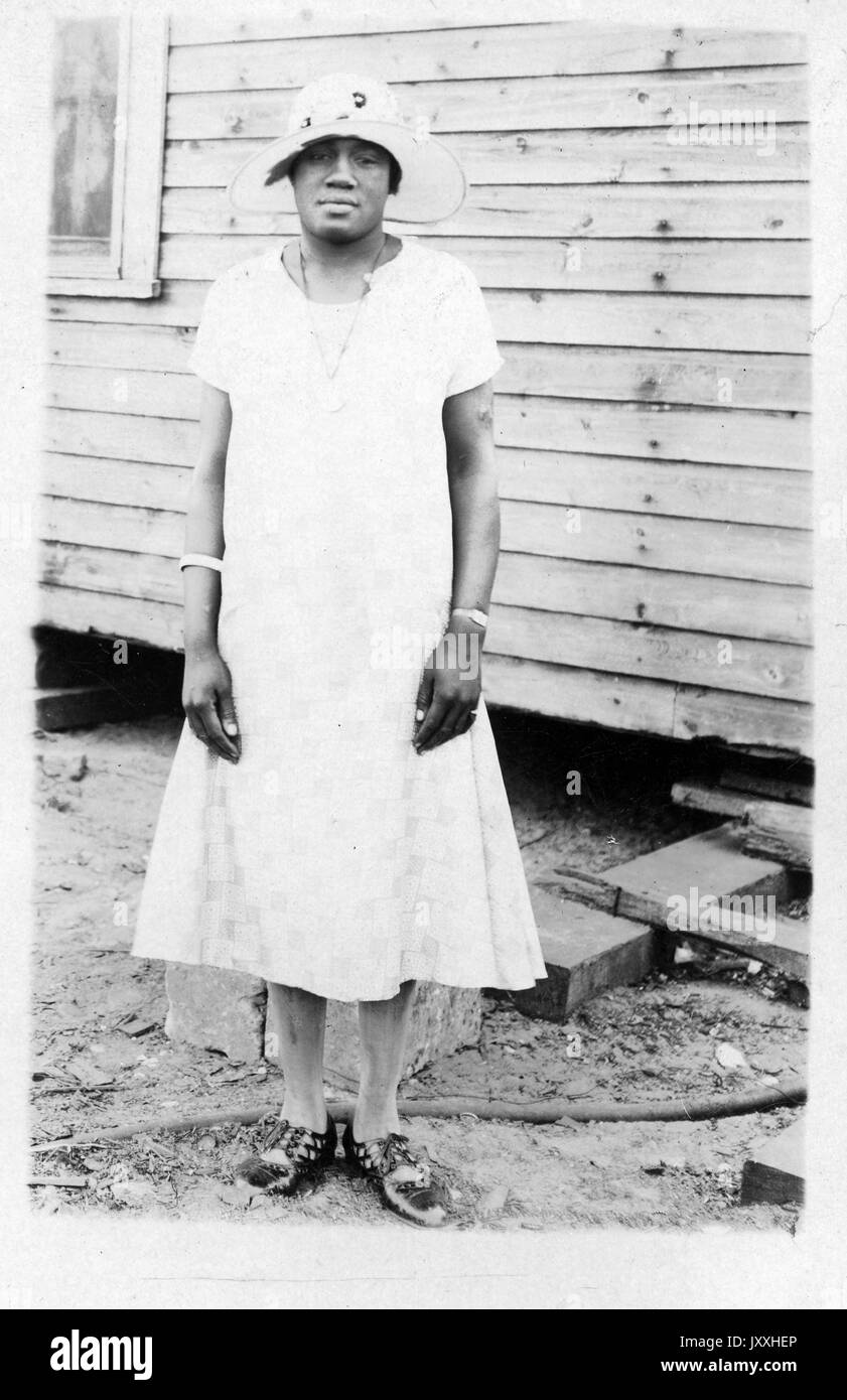 Portrait debout de femme afro-américaine mature, portant robe légère, collier, bracelets et chapeau, debout devant la maison, expression neutre, regarder vers le bas, 1920. Banque D'Images