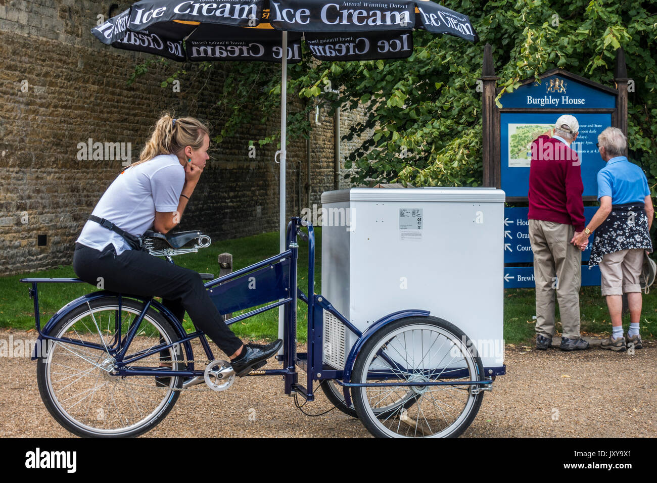 Une jolie fille vendeur de crème glacée, assis sur un vélo, à regarder un vieux couple, à l'extérieur de l'entrée des visiteurs à Burghley House, près de Stamford, England, UK. Banque D'Images