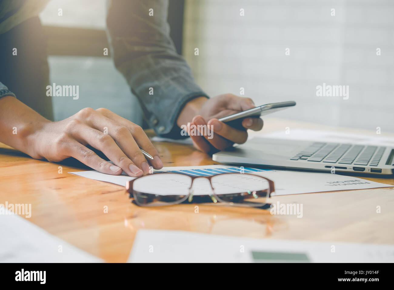 Business man holding smartphone et de l'écriture papier financière et de travail de l'ordinateur portable sur un bureau en bois bureau en intérieur avec la lumière du matin. Vintage effet de filtre. Banque D'Images