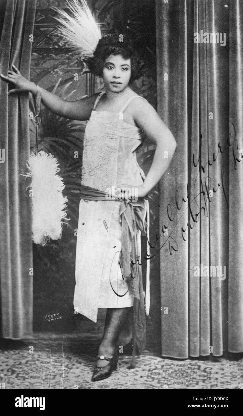 Portrait d'une femme afro-américaine debout et penchée avec son bras droit sur un rideau, elle porte une robe de couleur claire avec une écharpe autour de sa taille, son bras gauche repose sur sa hanche, elle a une grande décoration de plumes dans ses cheveux, 1920. Banque D'Images