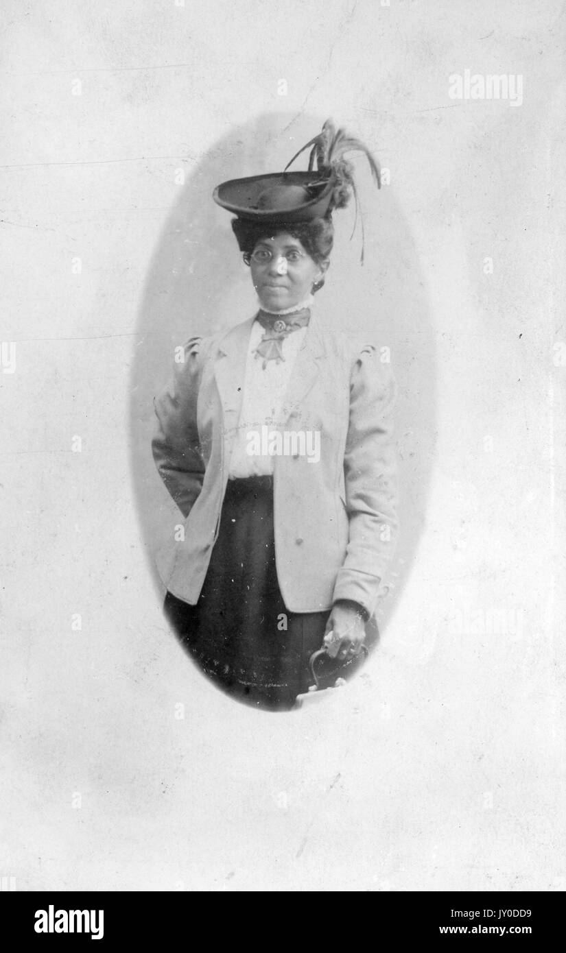 Portrait d'une femme afro-américaine debout tenant un sac à main dans sa main gauche, elle porte une jupe de couleur foncée et un chemisier et une veste de couleur claire, elle porte des lunettes et un chapeau très décoré, 1930. Banque D'Images