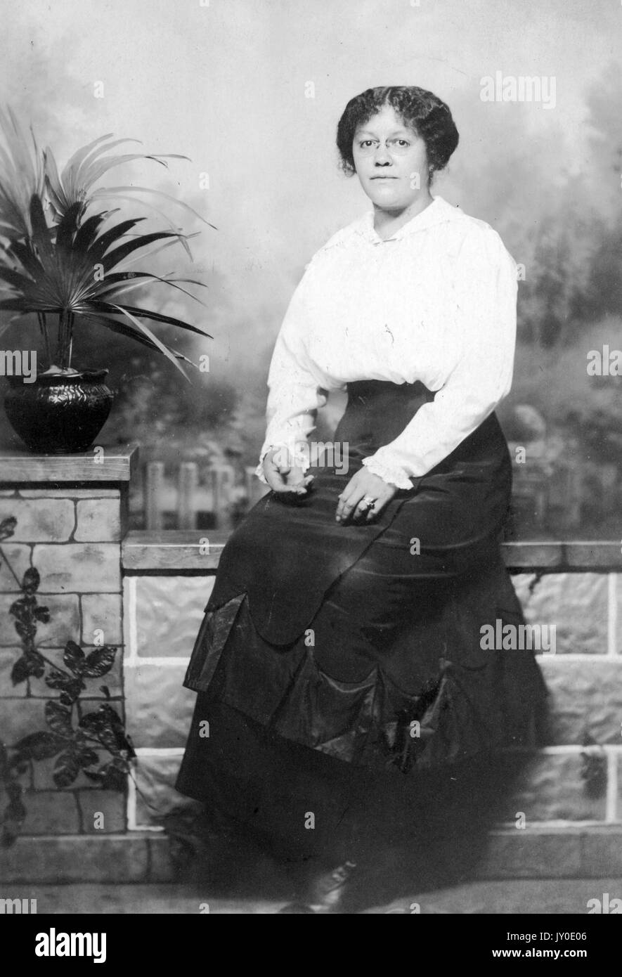 Portrait d'une femme afro-américaine assise devant une murale sur un banc avec un motif de fausse brique, à gauche est un pot avec une plante à l'intérieur, elle porte une jupe longue sombre et un chemisier de couleur claire, elle porte des lunettes et ses mains sont sur ses genoux, 1915. Banque D'Images