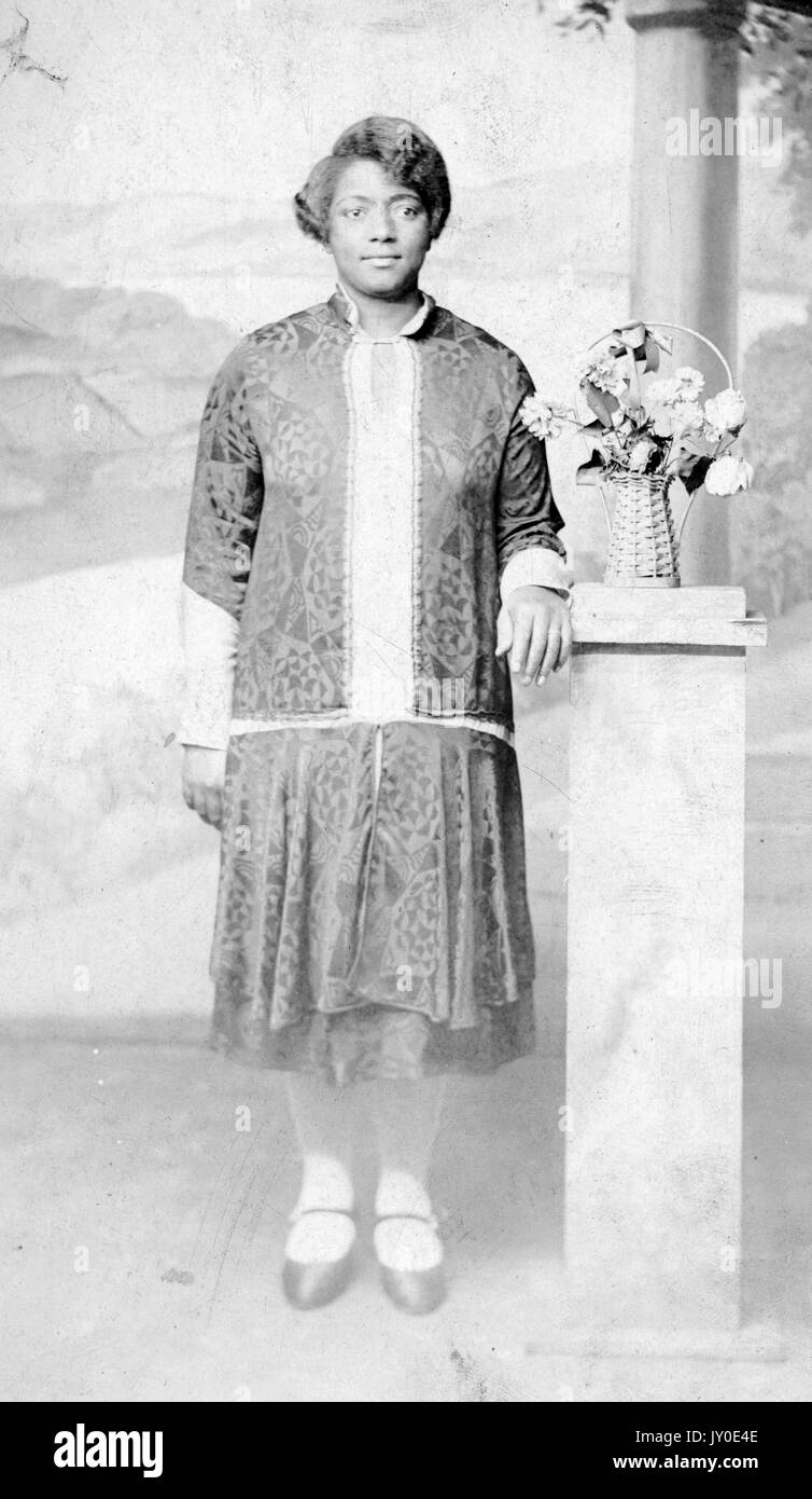 Portrait d'une femme afro-américaine debout devant une murale et à côté d'une longue prop tenant un bouquet de fleurs, elle porte une jupe et un chandail assorti avec un chemisier de couleur claire en dessous, un bras est à côté et l'autre repose sur le prop à côté des fleurs, 1925. Banque D'Images