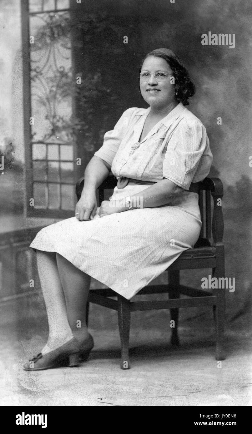 Portrait d'une femme afro-américaine souriante et assise sur une chaise en bois devant une murale, elle porte une robe longueur genou avec une ceinture et des talons, elle porte des lunettes, 1929. Banque D'Images