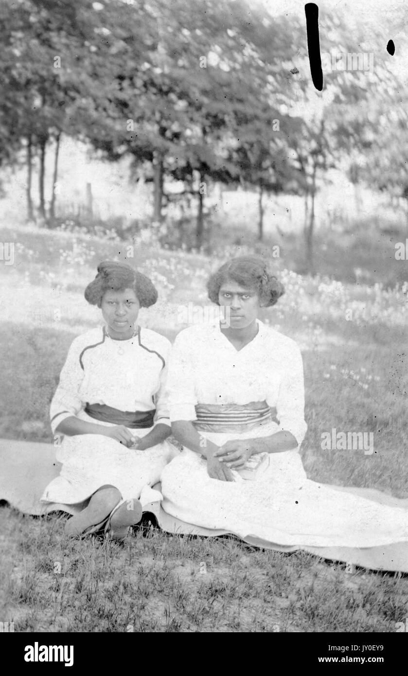 Portrait assis sur toute la longueur de deux jeunes Afro-américaines avec des expressions sérieuses à l'extérieur dans un champ, portant des robes légèrement colorées avec des accents sombres, 1920. Banque D'Images