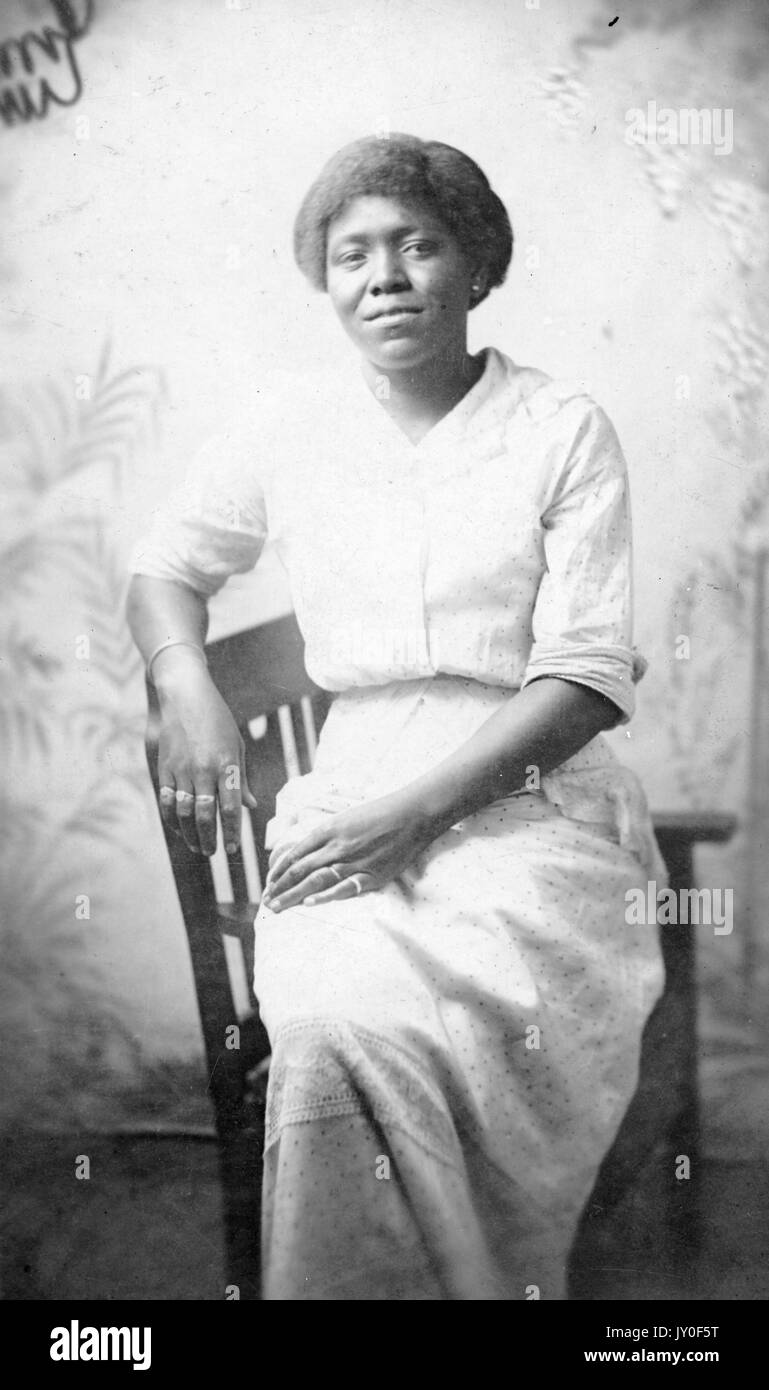 Portrait assis de trois quarts de longueur d'une femme afro-américaine mûre devant un fond peint, portant une robe à motifs, des boucles d'oreilles et des bagues de couleur légèrement, 1925. Banque D'Images