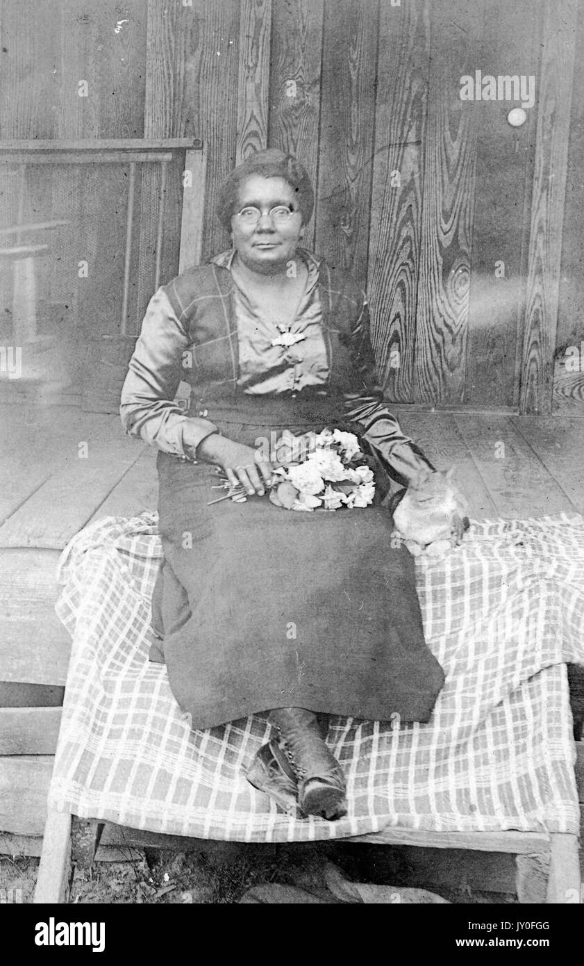 Portrait assis sur toute la longueur d'une femme afro-américaine mûre à expression neutre assise sur une couverture à motifs devant un bâtiment, portant un chemisier sombre, une jupe, et une bottes et tenant un bouquet de fleurs, 1925. Banque D'Images