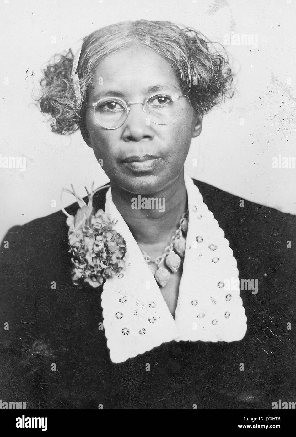 Tête de femme afro-américaine mature, portant un chemisier sombre avec collier clair, collier, broche de fleur, lunettes et barrettes de cheveux, expression neutre, 1925. Banque D'Images