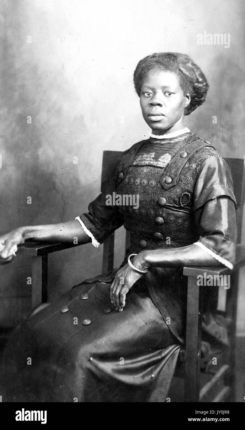 Portrait d'une femme afro-américaine assise dans une chaise en bois, ses mains sont placées sur les bras de la chaise et elle porte une longue robe de couleur foncée avec quelques garnitures de couleur claire, 1915. Banque D'Images