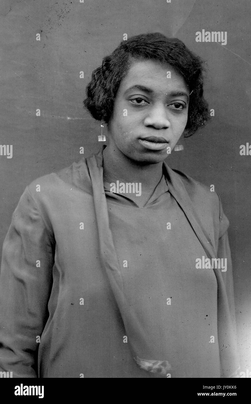 Tête de femme afro-américaine mature, portant un chemisier de couleur foncée, une écharpe et des boucles d'oreilles de couleur foncée, expression neutre, 1929. Banque D'Images