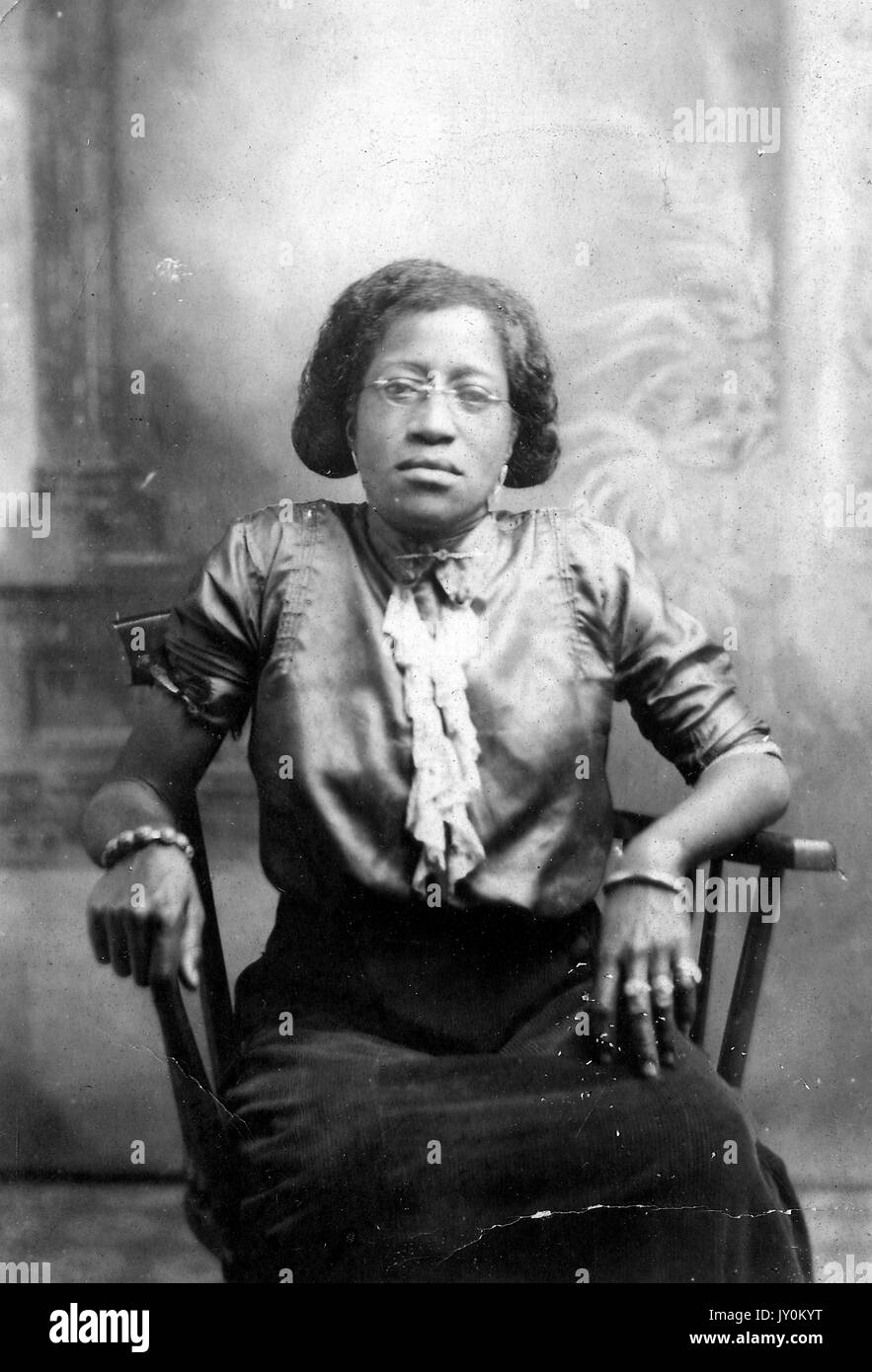 Trois quarts de longueur portrait assis de femme afro-américaine mature, portant un chemisier clair, jupe sombre, bijoux et lunettes, expression neutre, 1915. Banque D'Images