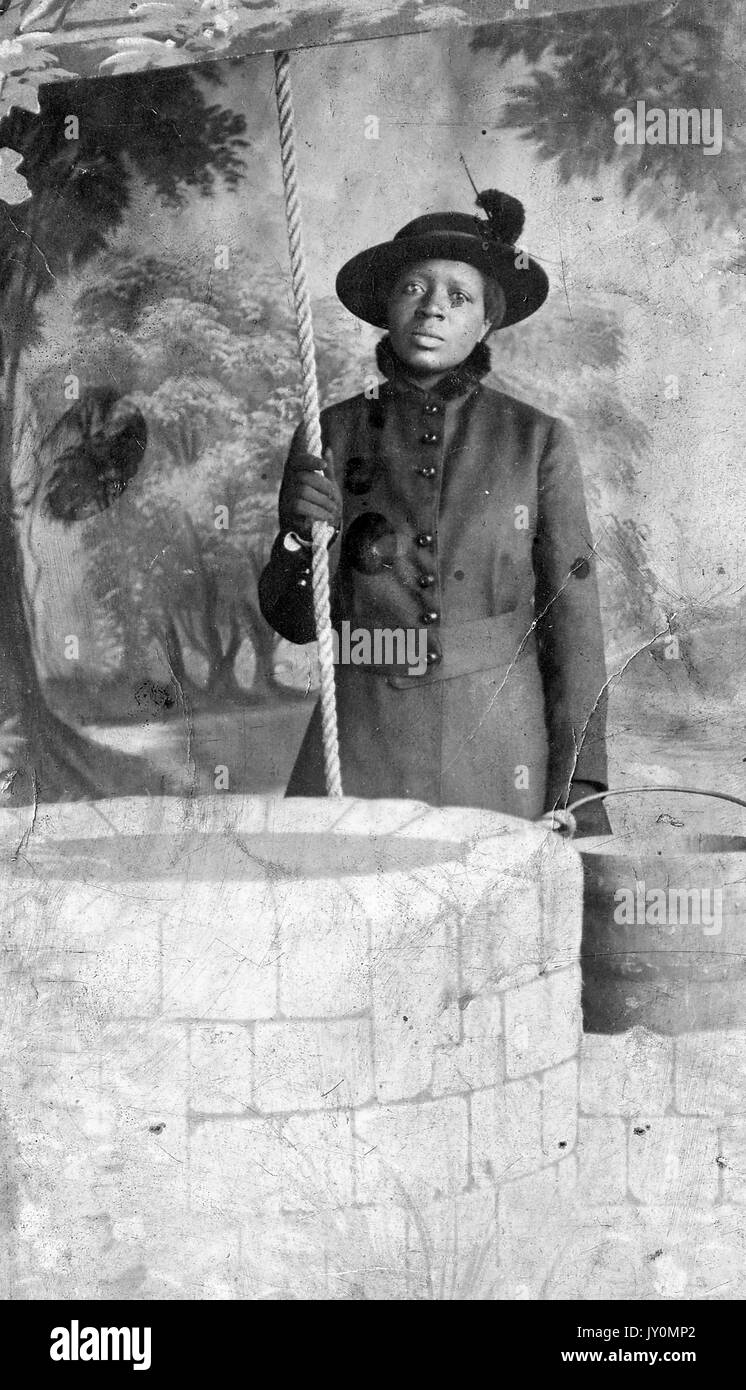 Portrait de trois quarts de femme afro-américaine, debout derrière un faux fond bien et devant une forêt de toile de fond, tenant une corde dans sa main droite, portant une robe sombre et un chapeau sombre, avec une expression faciale sérieuse, 1920. Banque D'Images