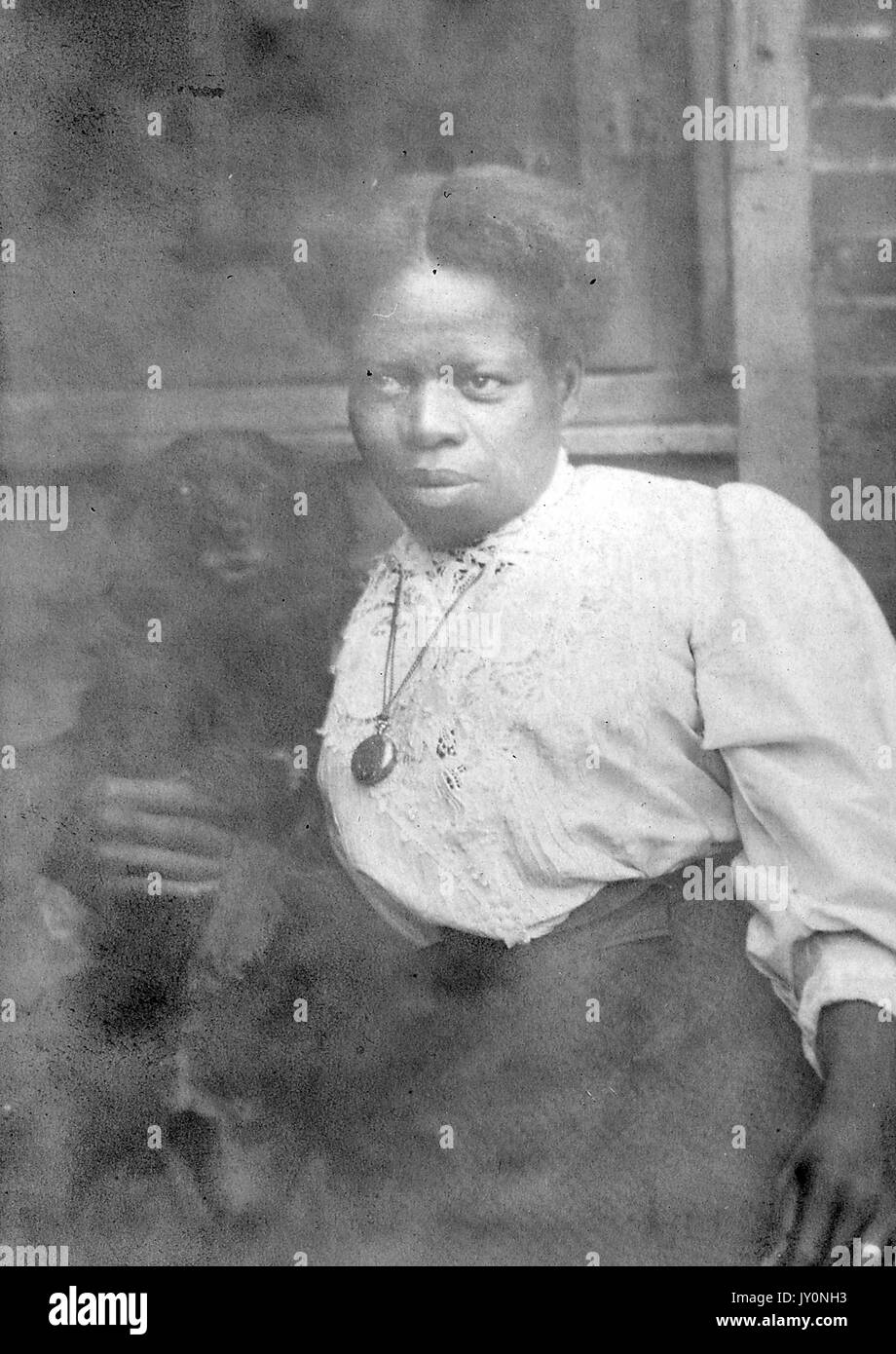 Portrait de trois quarts d'une femme afro-américaine devant un mur et une fenêtre en briques, portant un chemisier blanc et une robe sombre, portant un pendentif autour de son cou, avec une expression faciale sérieuse, 1920. Banque D'Images